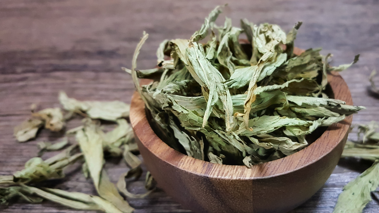 A wooden bowl, filled with dried stevia leaves, sits gracefully atop a wooden table, offering a natural touch to the scene. Nearby, scattered on the same table, more dried stevia leaves evoke a sense of harvest and herbal abundance.