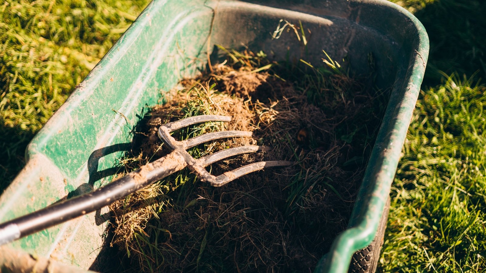 A pitchfork sits on a green wheelbarrow filled with straw and organic matter.