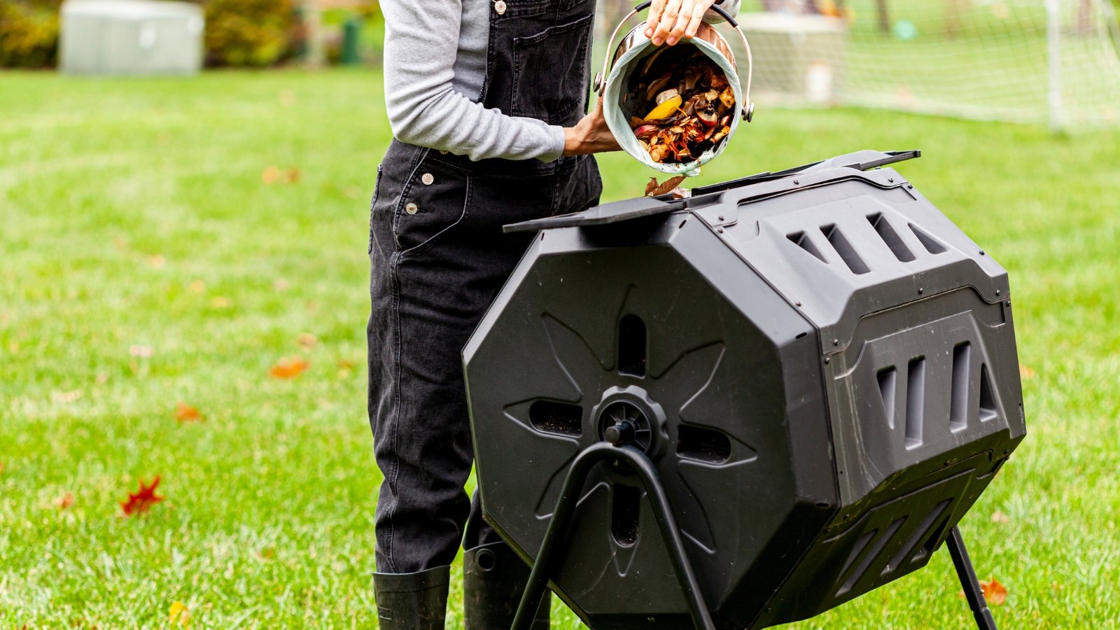 The image shows a woman tilting a small plastic container to release a mixture of vegetable scraps and other biodegradable waste into a larger container intended to enrich the soil.