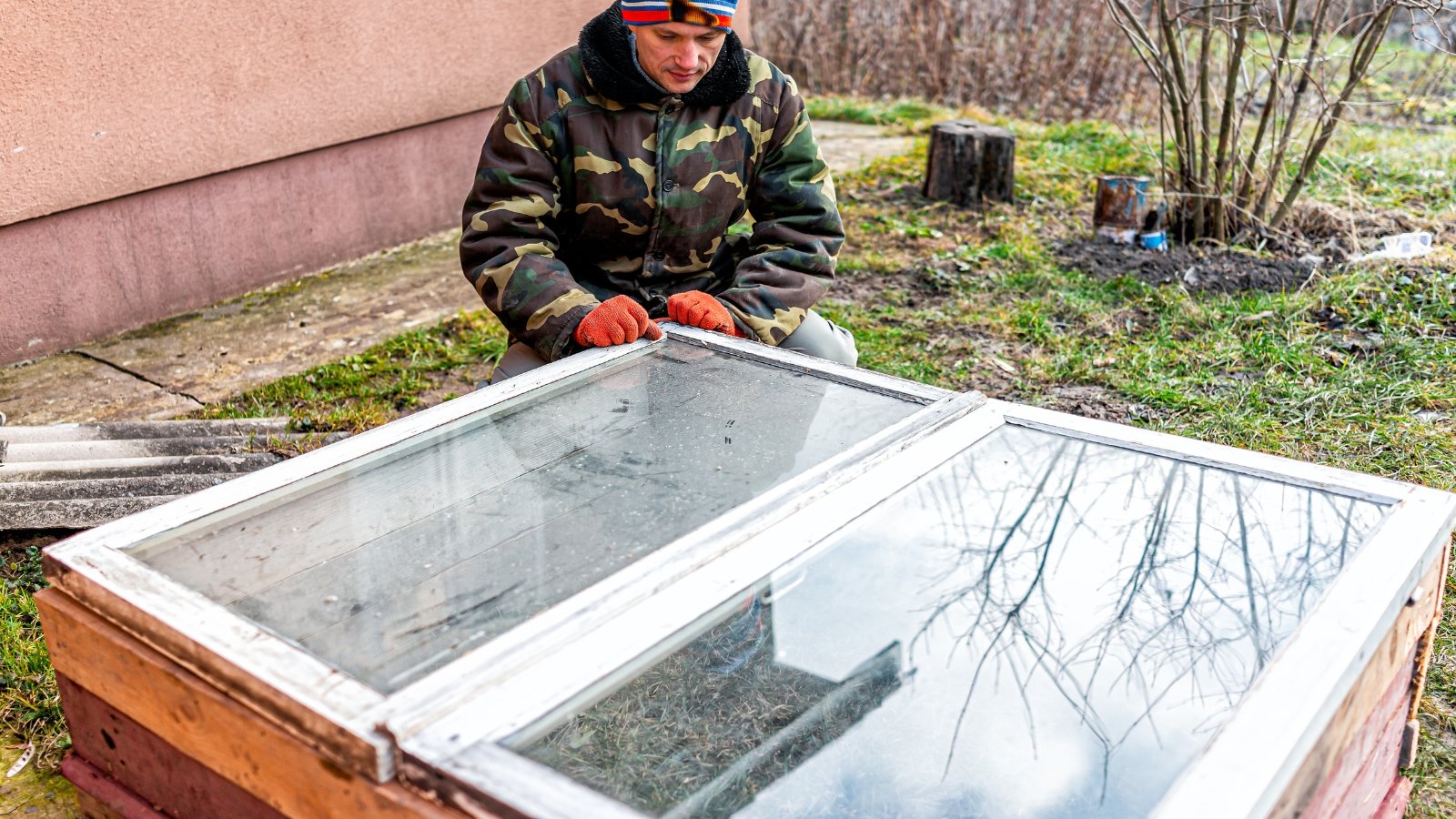 A person in a green jacket building a cold frame from repurposed windows, kneeling beside a raised garden bed.