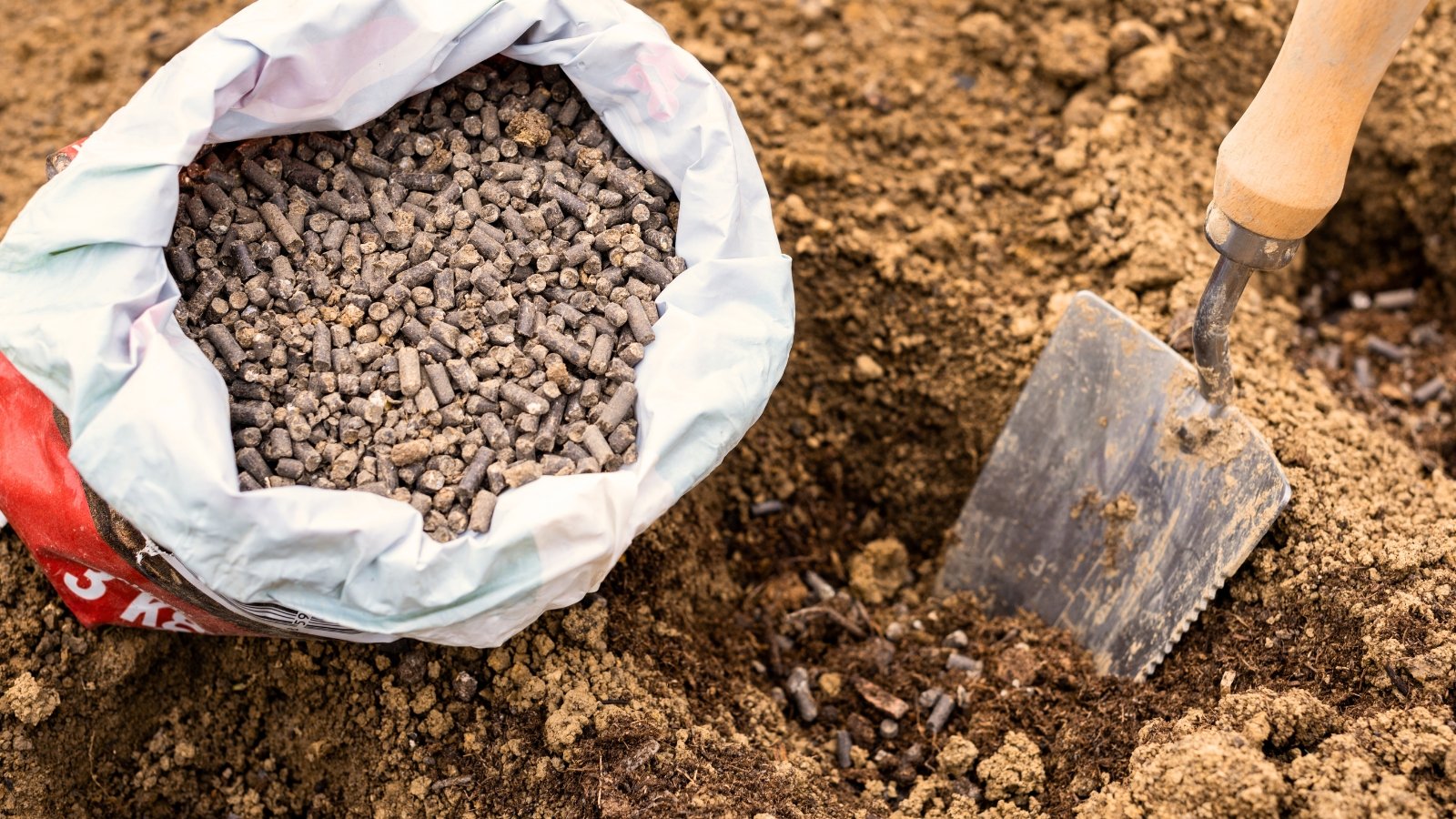 A bag of compressed chicken manure pellets rests on the ground next to a small garden trowel embedded in the dirt.