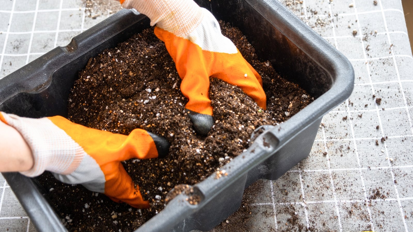 Close-up of a gardener's hands in orange gloves mixing the soil mixture in a large black container.