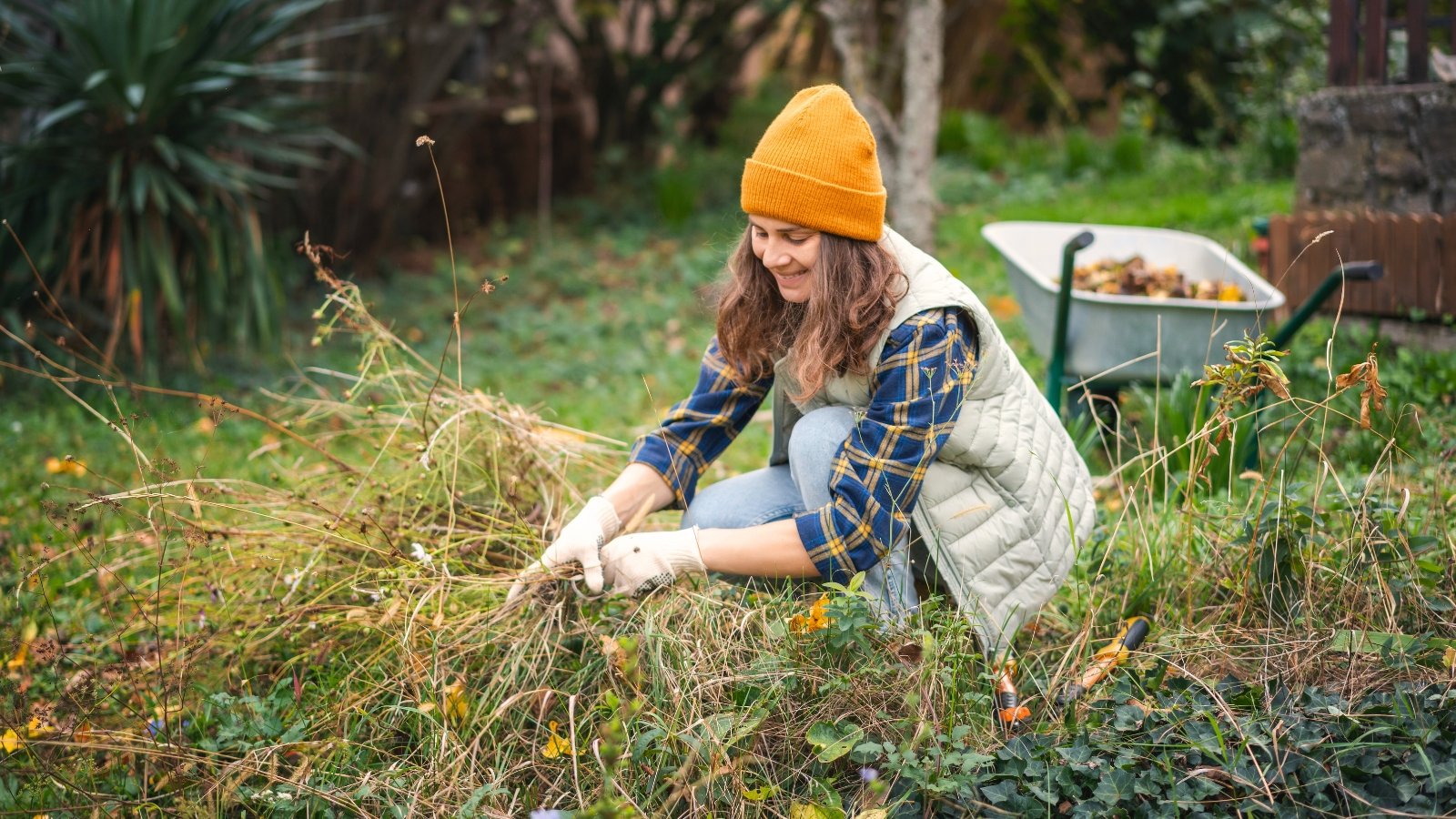 A woman in a yellow hat and checkered shirt clears dried plants from the autumn garden, surrounded by fading foliage and seasonal blooms.
