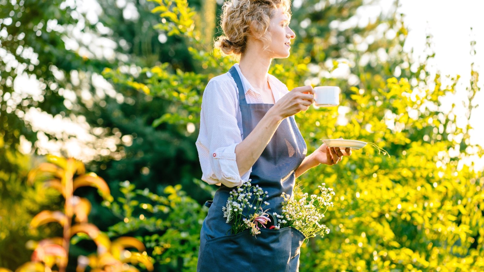 Beautiful portrait of a thoughtful blond curly woman with wildflowers in her pocket, holding a cup of coffee in a sunny garden. The woman is wearing a white shirt and a blue apron.